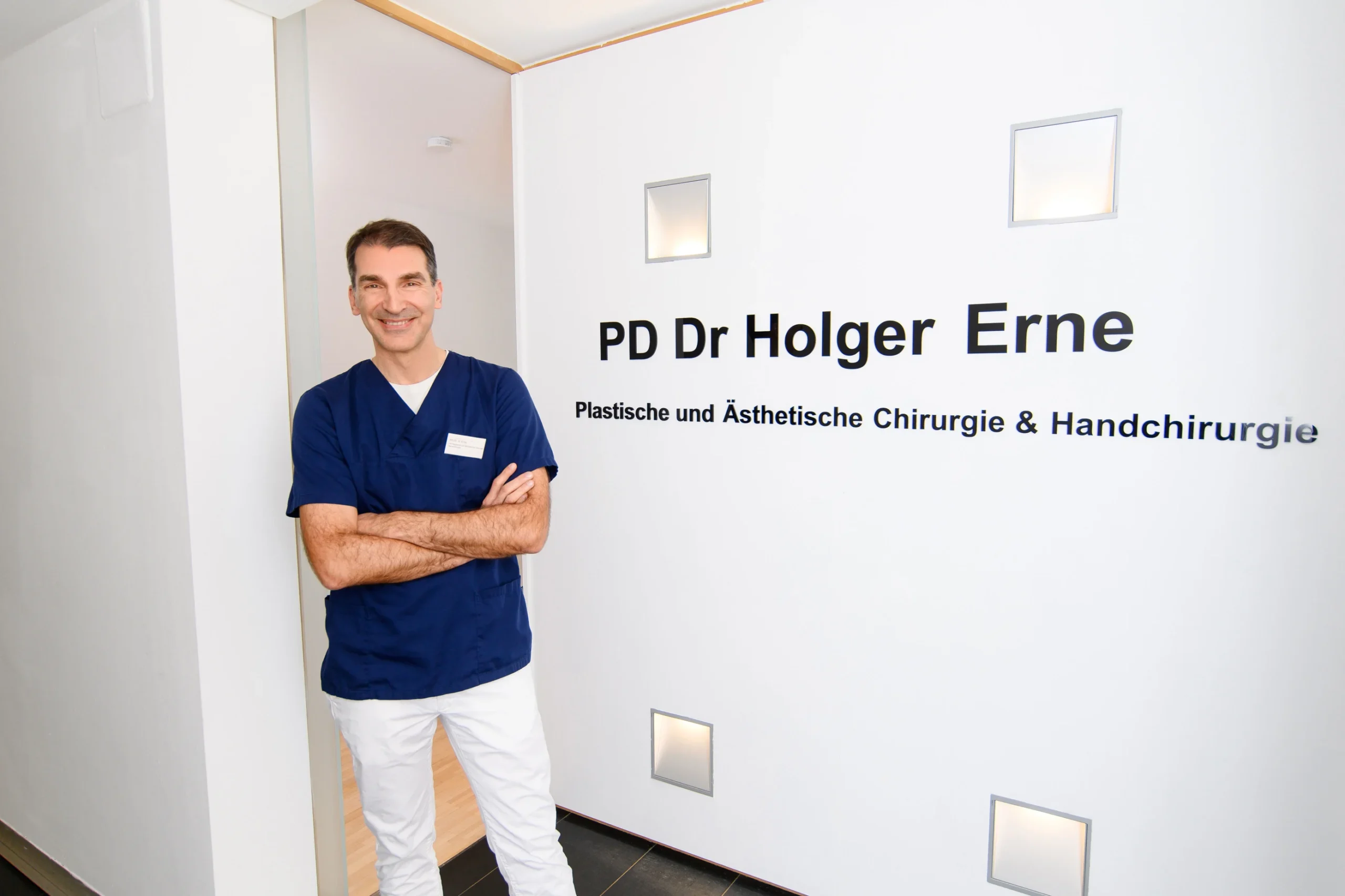 Zu sehen ist Dr. Holger Erne sympathisch in die Kamera lächelnd. Er steht vor seinem Patientenzimmer, am Eingang stehen große Buchstaben an der Wand mit seinem Namen 